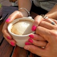 A woman with pink nails holding a cup of coffee while her husband holds her left hand.