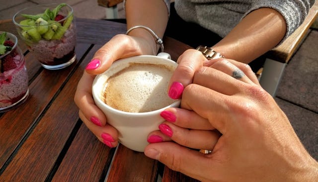 A woman with pink nails holding a cup of coffee while her husband holds her left hand.