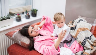 Mother wearing a pink robe fighting a stomach bug with her blonde child wearing a white shirt sittin...