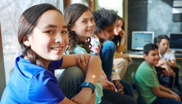 A tween girl with a braid in a blue shirt smiling while sitting next to a group of students in the b...