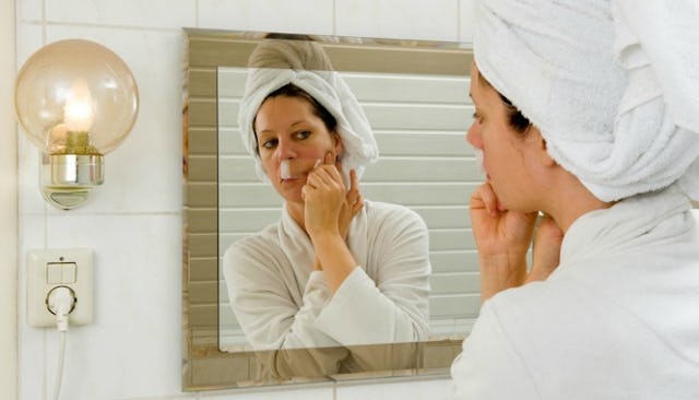A woman taking the chin hair off her face in a mirror wearing a white bathrobe and white towel on he...