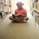A Black girl sits cross-legged reading a book on the floor of a library, in between rows of bookcase...