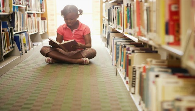 A Black girl sits cross-legged reading a book on the floor of a library, in between rows of bookcase...