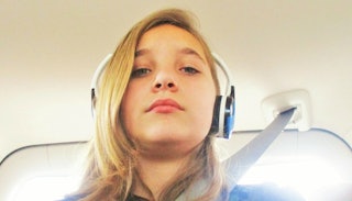 A teenager taking a selfie with her buckled up in the car while wearing a blue shirt and blue and wh...