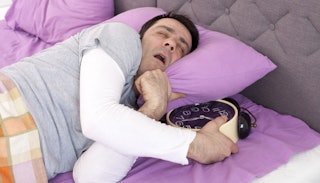 A sleeping husband in a grey shirt with white sleeves sleeping in purple bed sheets while holding hi...