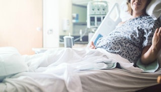 A pregnant woman wearing a patient gown in a hospital bed with medical devices in the background