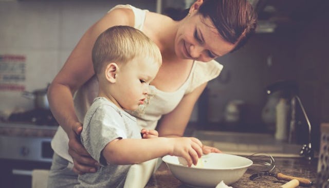 A mother standing in the kitchen and cooking with her son who is in the phase between a baby and a b...