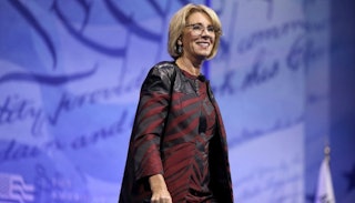 Betsy DeVos wearing a long-sleeved red and black brocade coat with a blue background on stage
