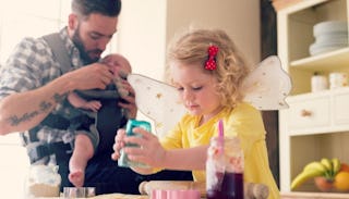 A young father feeding his baby while his daughter is playing next to him in the kitchen.