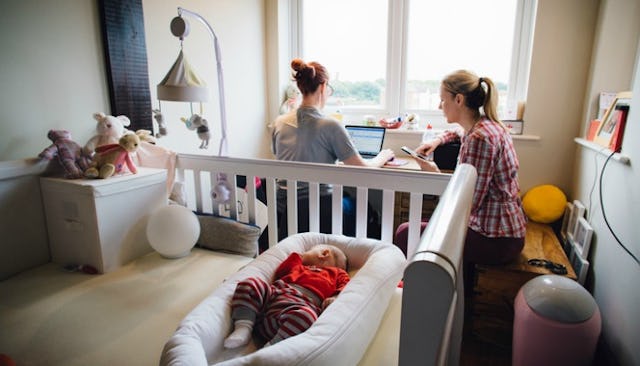 Two young lesbian moms sitting next to each other while their baby peacefully rests in a crib just b...