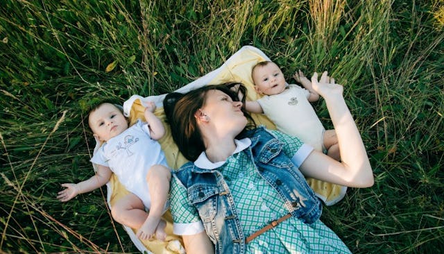 A mother in a blue dress with a denim vest, lying on the yellow blanket in the grass with her twin b...
