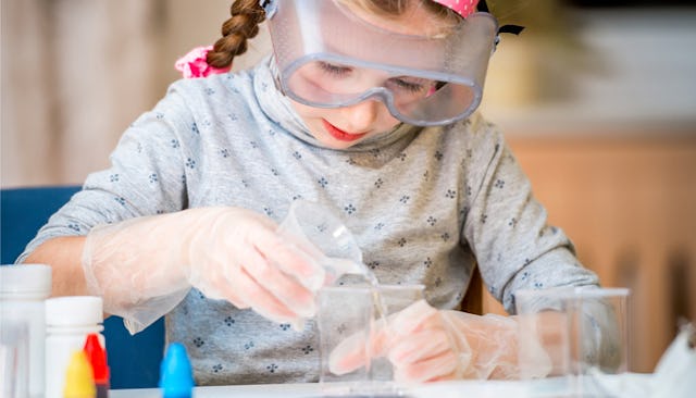 A girl wearing protective goggles in a gray shirt and plastic gloves preparing her science fair proj...