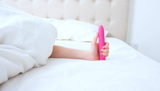 A woman lying in her bed and holding a pink sex toy in her hand while covered with a white duvet