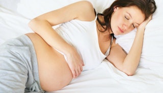 A pregnant brunette woman sleeping on her side in white bed sheets wearing a white tank top and grey...