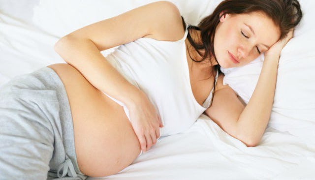 A pregnant brunette woman sleeping on her side in white bedsheets wearing a white tank top and gray ...