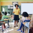 A teacher in a yellow sweater, black skirt, and black shoes with open arms talking to children seate...