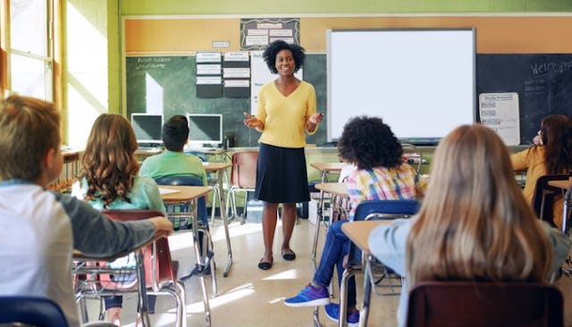 A teacher in a yellow sweater, black skirt, and black shoes with open arms talking to children seate...