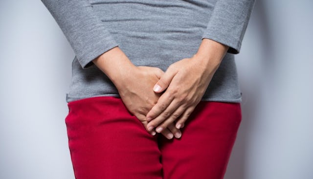 A woman standing in a gray shirt and red pants while covering her private area.