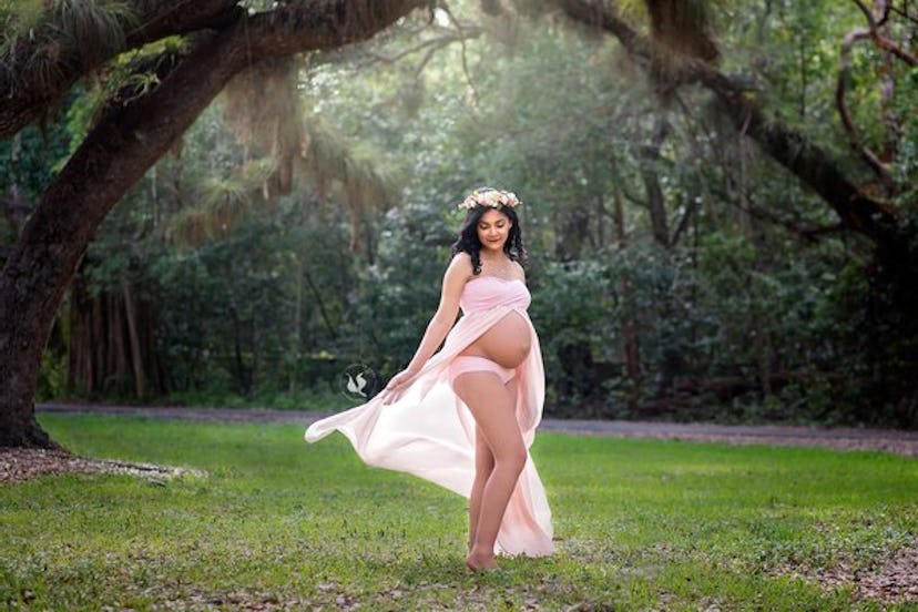 A pregnant woman posing outdoors in a forest wearing a cut-out pink dress that’s revealing her belly