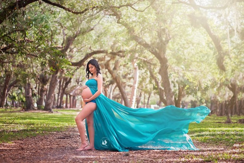 A pregnant woman caressing her belly surrounded by trees while her teal dress is floating behind her...