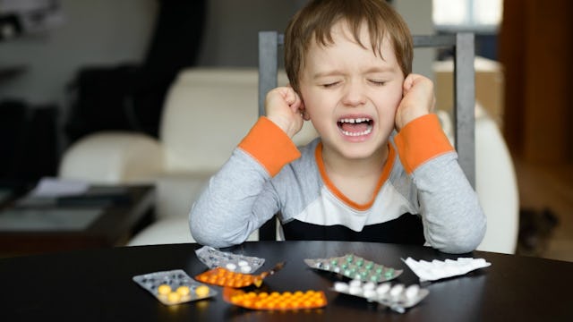 A kid is yelling and placing his hands on ears while set of ADHD medication are in front of him on t...