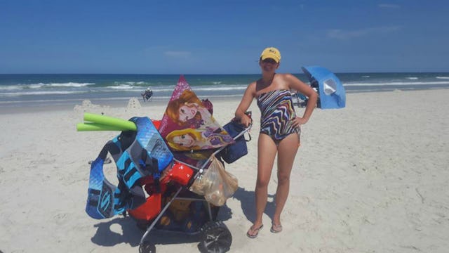 A woman posing next to a baby stroller filled with beach gear with the sea in the background