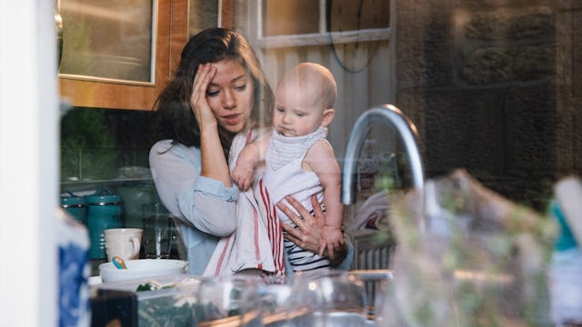 Woman Holding Her Head And Standing In The Kitchen With A Baby