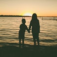 A taller girl and a shorter boy holding hands slightly darkened, watching the sunset with the sea in...