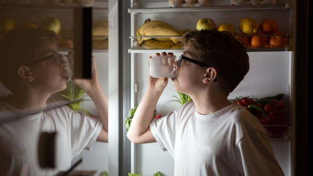 A boy in a white shirt drinking a glass of milk in front of an open refrigerator at a late hour as h...