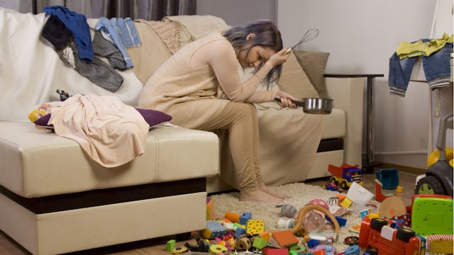 A mother sitting on a couch, holding her head, overwhelmed by the chaos in her house