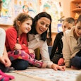 A brunette, short-haired preschool teacher surrounded by six children showing them some drawings on ...