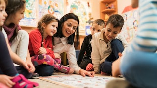 A brunette, short-haired preschool teacher surrounded by six children showing them some drawings on ...