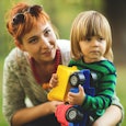 A babysitter crouches next to a toddler holding a plastic truck toy in the park, and both look to th...