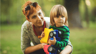 A babysitter crouching next to a toddler, who is holding a plastic truck toy in a park