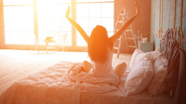 A happily divorced woman stretching in her bed with her arms raised in a sunlit bedroom
