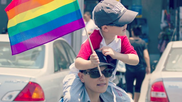 A small boy holding an LGBTQ flag while sitting on his parents shoulders
