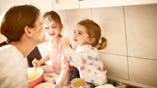 Two girls sitting and smiling on a kitchen countertop while covering their mom-s face with flour