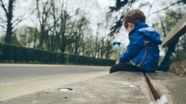 A sad little boy with autism in a blue jacket sitting alone on a bench
