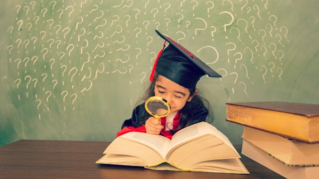 A preschool child sitting in front of a chalkboard full of question marks looking at a book through ...