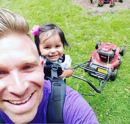 A dad carrying his daughter in a baby carrier while mowing the grass 