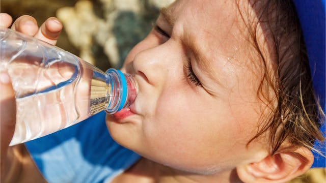 A boy in a blue shirt in the sun drinking water from a bottle