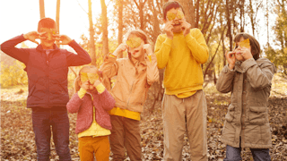 Five children in a forest wearing clothes in a brown, mustard and beige palette, holding yellow leav...
