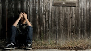 A desperate boy sitting on the ground while leaning against the wall and covering his face with his ...