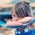 A shy young girl wearing blue clothes is leaning with her arms and head on the blue swing in a park 