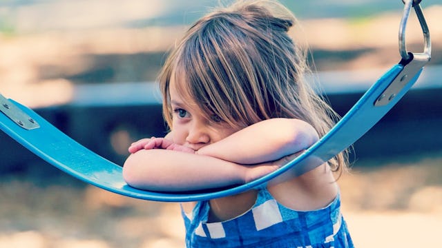 A shy young girl wearing blue clothes leaning with her arms and head on the blue swing in a park