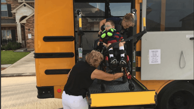 Two people assisting a disabled child in a wheelchair entering a school bus on a disability-speciali...