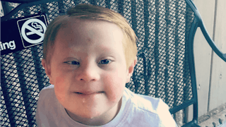 A child with Down Syndrome sitting on a blue chair directly facing the camera and smiling in a white...