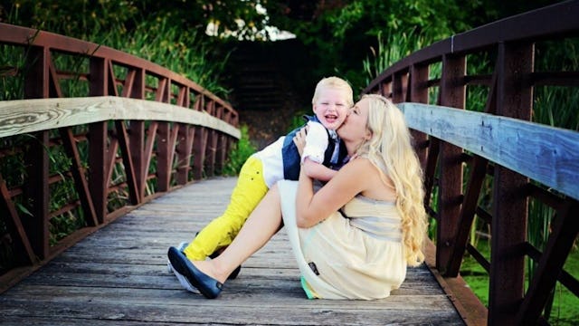 A mother sitting on a wooden bridge holding her child and kissing it on the cheek