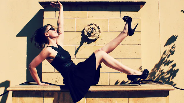 A woman in a black dress semi-lying with one leg and arm lifted outside under direct sunlight during...