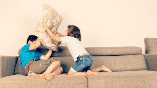Two siblings fighting on a grey couch and the girl is using a pillow to hit her brother